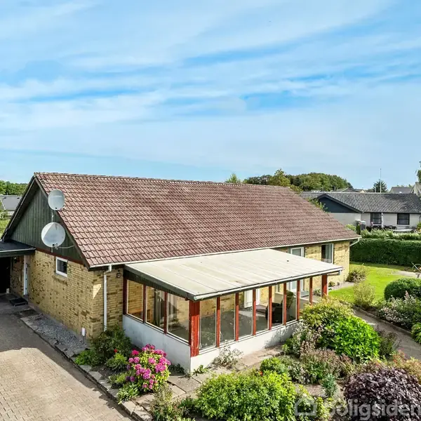 Et gulstenshus med en veranda ud mod en have fyldt med buske og blomster; en brolagt indkørsel forbinder huset med en garage. Husene i baggrunden er omgivet af træer.
