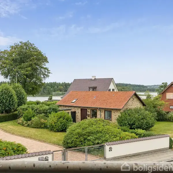 Et lille hus med rødt tegltag står omgivet af frodig have. I baggrunden ses flere huse, en sø og træer, under en klar blå himmel.