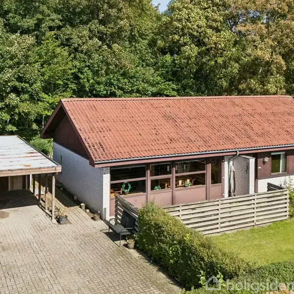 Et enfamiliehus med rødt tegltag og en carport står omgivet af grønne træer. Forpladsen er brolagt med sten, og der er en lav hæk foran huset.