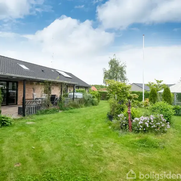 Et hus med skråt tag står stille i en frodig have. Omkring det er græsplæne, buske og et lysthus. Skyer ses på den blå himmel.