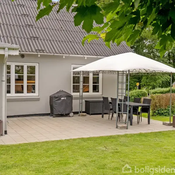 Terrasse med bord og stole under en hvid parasol. En grill står ved siden af huset med gråt tag. Græsplæne omgiver området, med buske og et andet hus i baggrunden.