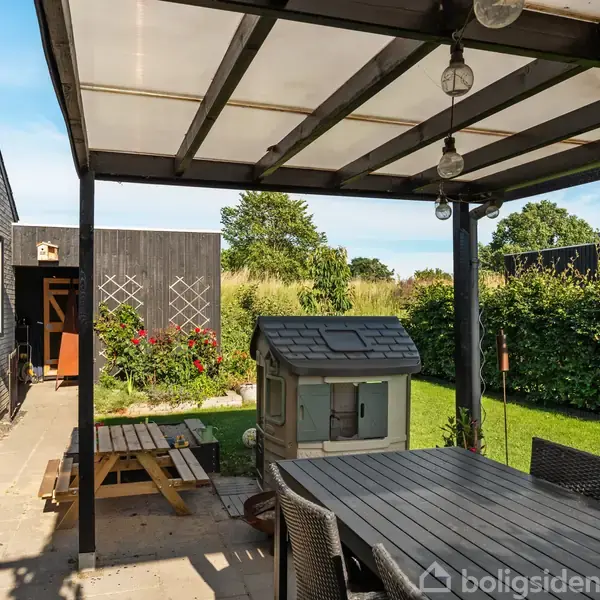 En overdækket terrasse med spisebord og stole står stille under et klart tag. Baggrunden viser en grøn have med buske og blomster i et solrigt vejr.