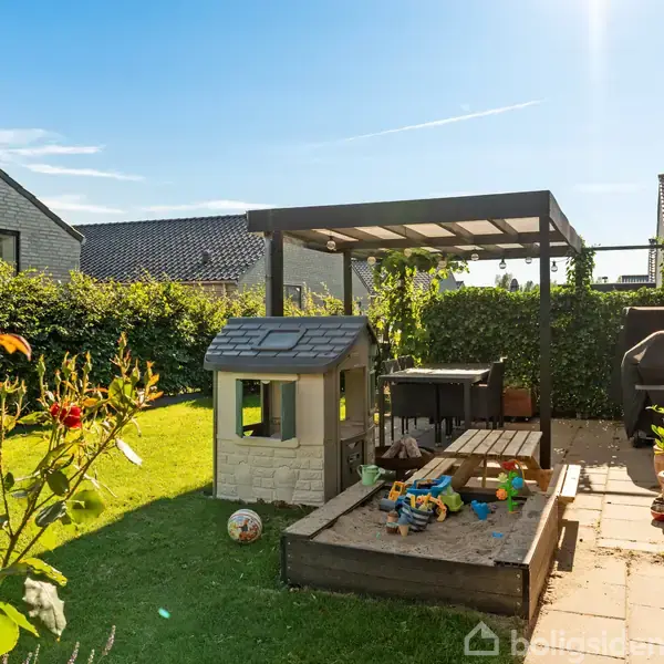 Et lille legehus står på en græsplæne omgivet af et hegn. Ved siden af er en pergola med klatrestativer og legetøj. Solen skinner fra en klar blå himmel.