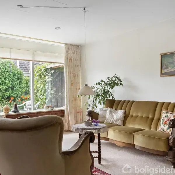 En stue med vintage møbler, herunder en sofa og to lænestole, placeret på et rødt tæppe. Store vinduer giver udsigt til en frodig have. En lampe hænger fra loftet.