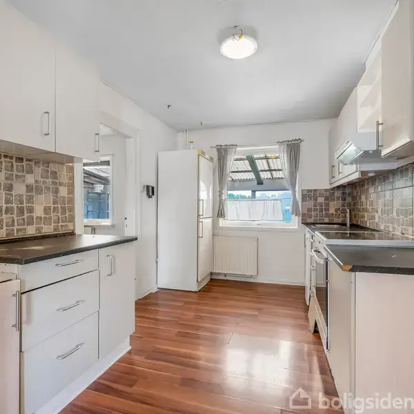 Et lyst køkken med hvide skabe og mørkebrune trægulve. Komfur og opvaskemaskine står langs væggen. Fliser med naturinspireret mønster pryder væggen, og lyset strømmer ind fra vinduerne.