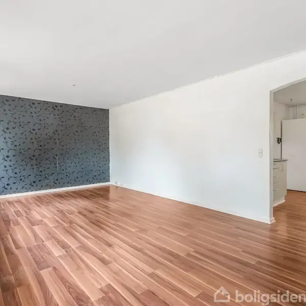 Et tomt rum med trægulv strækker sig mod en mørk accentvæg med mønster. Til højre forbinder en buegang rummet til et køkken med hvide skabe og et vindue.