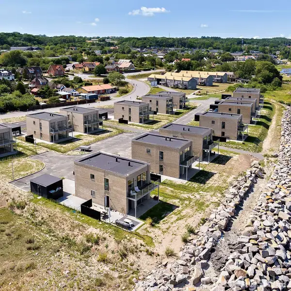 Moderne rækkehuse står stille langs en kyststrækning omgivet af en stenmur. Bagved ses en by med spredte huse og grønne områder under en klar himmel.