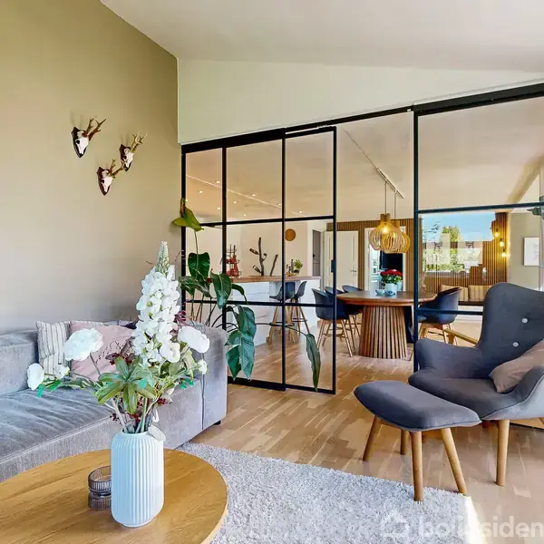 Stue med grå sofa og blomster i vase på rundt bord. Stor glasvæg adskiller spiseområde med træbord og hængelamper. Billede af barn hænger på væggen med dyredekorationer.