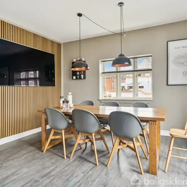 Et spisebord i træ står i midten af et moderne rum. Seks grå stole er placeret omkring bordet. På væggen hænger en stor fladskærm. En plakat med teksten "Old Trafford" ses ved siden af et vindue. To sorte pendellamper hænger over bordet.