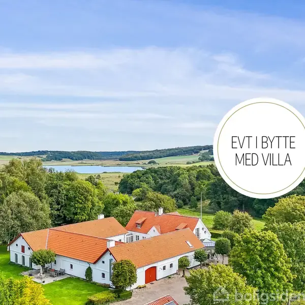 Gård med røde tage ligger i frodigt landskab nær en sø. Rund cirkeltekst siger: "EVT I BYTTE MED VILLA".