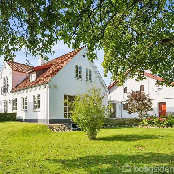 En hvid hus med rødt tag står i en frodig have omkranset af træer og buske. Grønt græs og blå himmel skaber en idyllisk scene.