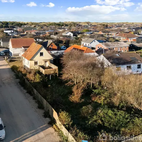 Et villakvarter ses fra oven, med huse i forskellige farver og former langs en vej. Omkringliggende haver og buske omgiver husene, mens blå himmel strækker sig i baggrunden.