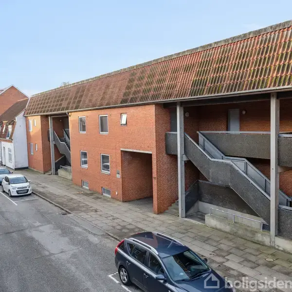 En række af røde murstensbygninger med tilhørende udendørs trapper står ved en stille gade med parkerede biler. Himlen er klar og solbeskinnet.