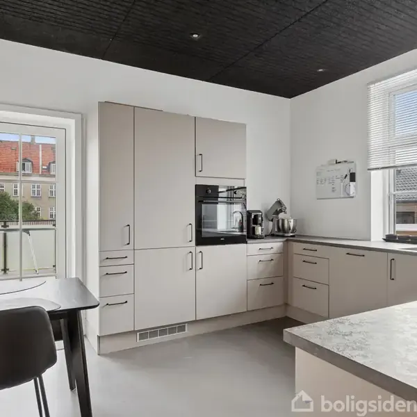 Et moderne køkken med beige skabe og integreret ovn, står ved siden af et lille spisebord og to stole. Store vinduer giver udsigt til omkringliggende bygninger.