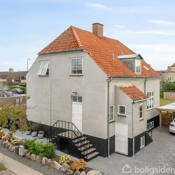 Et to-etagers hus med hvid facade og rødt tegltag står i en gårdhave. Huset har flere vinduer og en trappe op til hoveddøren. Baggrunden viser et nabolag.