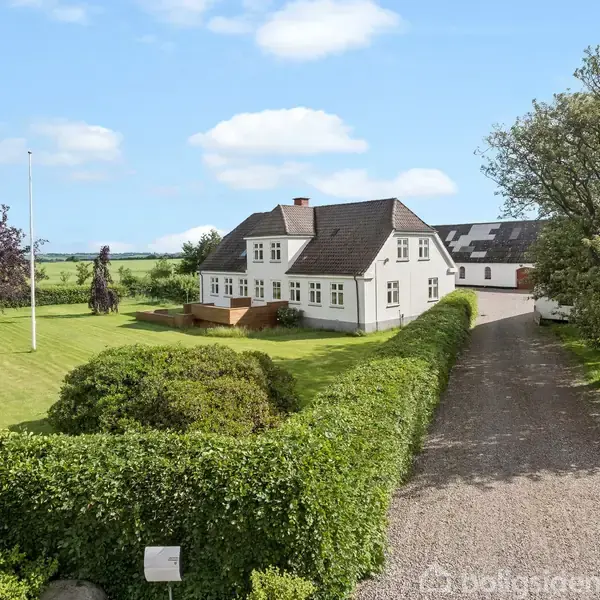 Hvidt to-etagers hus med mørkt tag står på en velplejet græsplæne omgivet af en hæk. En grusvej fører til huset. Området er landligt og åbent med marker i baggrunden.