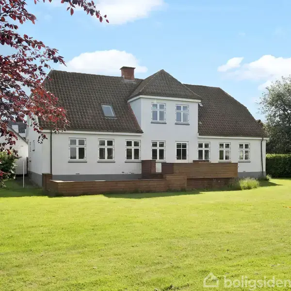 Stort hvidt hus med brunligt tag står i ro omgivet af en velplejet græsplæne og træer. Huset har flere vinduer og en terrassedækning.