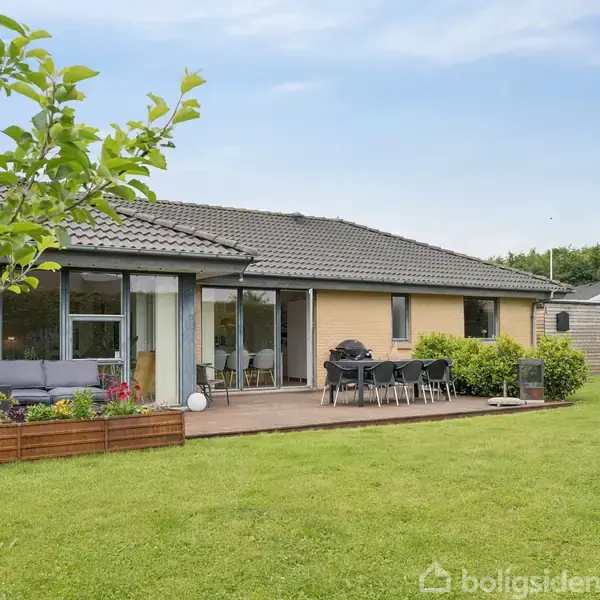 En étplans hus med brun facade står i centrum. En terrasse har havemøbler og grill. Huset ligger i en have med græsplæne og planter.