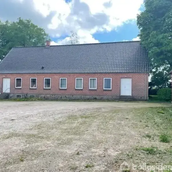 Et rødt murstenshus med fem vinduer står stille i en åben gårdsplads omgivet af grønne træer og græs på en solrig dag.