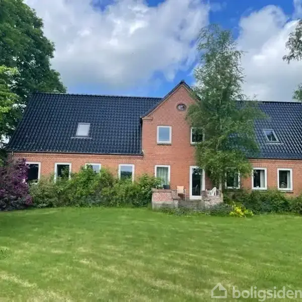 Et rødt murstenshus med sort tag står i en grøn have omkranset af frodigt løv, buske og træer under en delvis overskyet blå himmel.