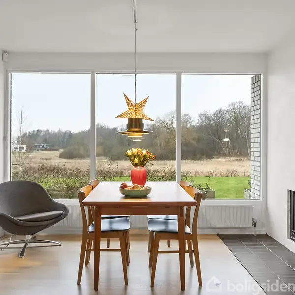 Et spisebord med blomster i en vase står foran store vinduer med udsigt til et åbent landskab. Ved siden af er der en grå lænestol og en tændt pejs.