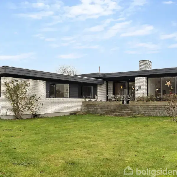 En hvid murstensvilla har store vinduer og en terrasse foran. Huset står på en grøn græsplæne med nogle buske i forgrunden under en blå, let overskyet himmel.