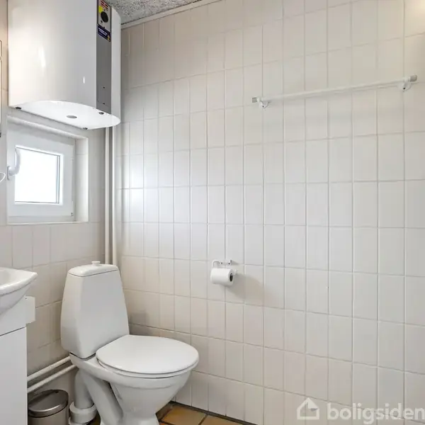 Toilet og vask i et flisebelagt badeværelse. Toilettet står ved siden af et lille vindue. Ovenfor er der en hvid vandvarmer. Der er et spejl og en håndvask med skab.