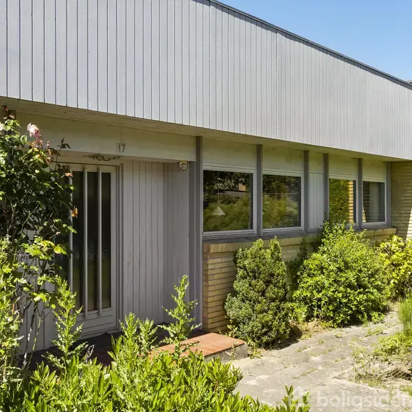 Et én-etagers hus med træbeklædning og store vinduer står omgivet af frodig vegetation i en have. Nummer 17 er synligt ved hoveddøren.