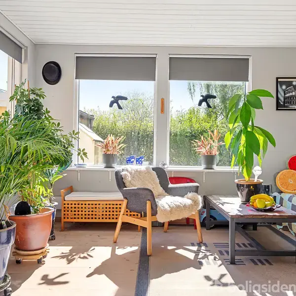 En solfyldt dagligstue med planter i krukker som hovedfokus. En behagelig lænestol med fåreskind står foran vinduer med udsigt til en have. En sofa med farverige puder er til højre.