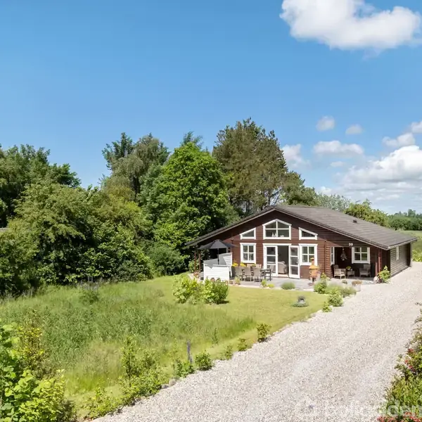 Hus set udefra med træbeklædning og store vinduer, stillestående på en grusvej omgivet af grønne marker og træer under en blå himmel med få skyer.