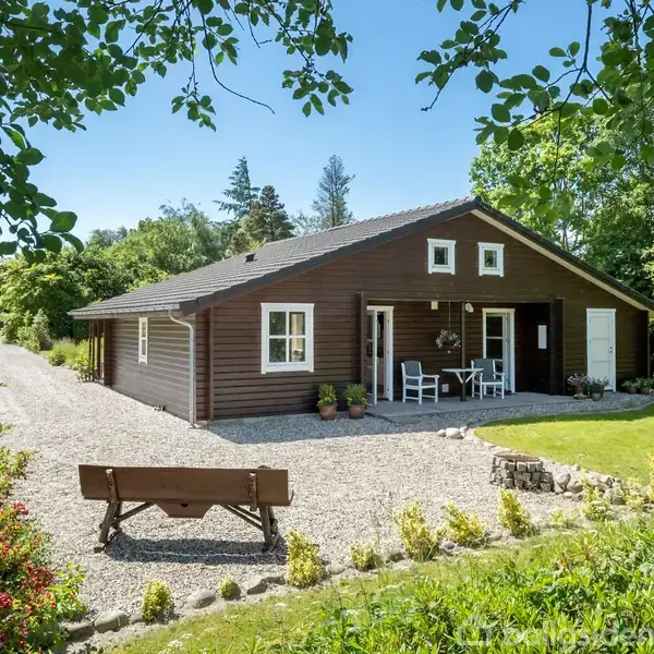 Et træhus med terrasse står omgivet af en grøn have. Foran huset ses en grusvej og en bænk. Sollys skinner ned gennem de tætte træer.