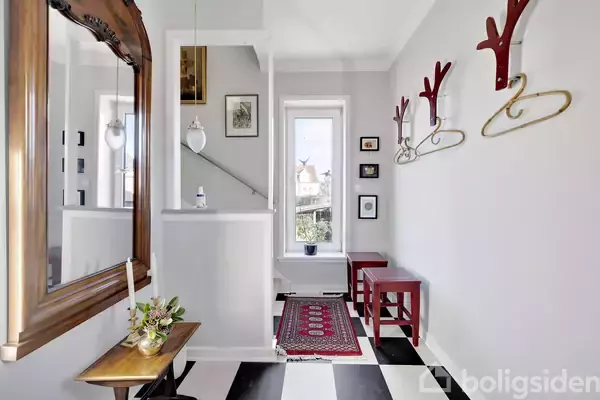 Spejl hænger på væggen. Skohylder og knagerækker pynter entréen med sort-hvidt ternet gulv. Et bord med planter står ved spejlet. Et smalt vindue viser udsigt udenfor.