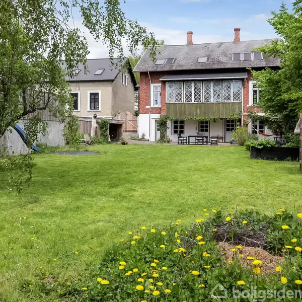 Hus med rød murstensfacade står for enden af en stor græsplæne med træer og blomster. En terrasse med borde og stole ses foran huset under en balkon.