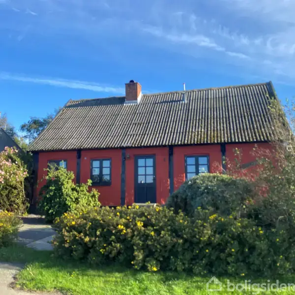 Et rødt hus med sorte vinduesrammer står omgivet af frodige buske og blomster i en have under en klar blå himmel.