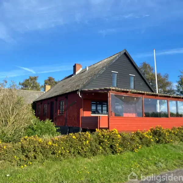 Rødt hus med glasindrammet veranda, beliggende på en grøn græsplæne omgivet af buske. Blå himmel og træer i baggrunden.