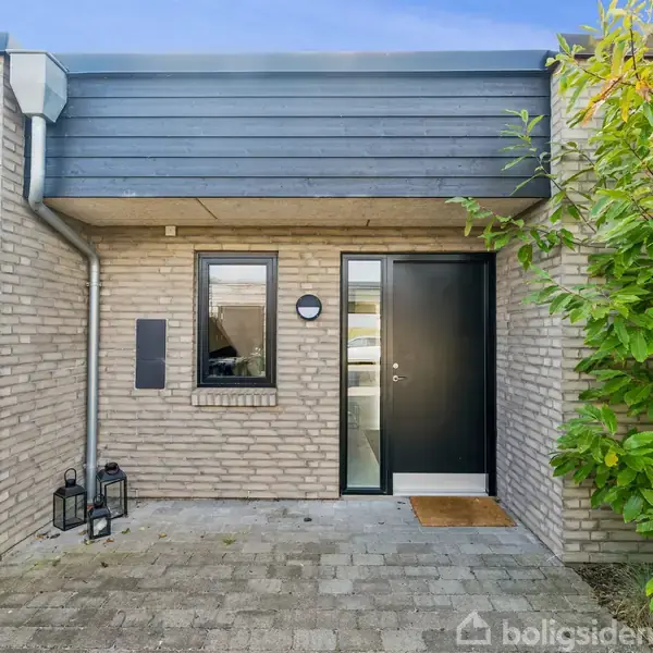 En moderne enfamilies hus har en mørk hoveddør ved siden af et vindue, omgivet af lyse mursten. To sorte lanterner står ved døren på stenbelægning, med grønne buske til højre.