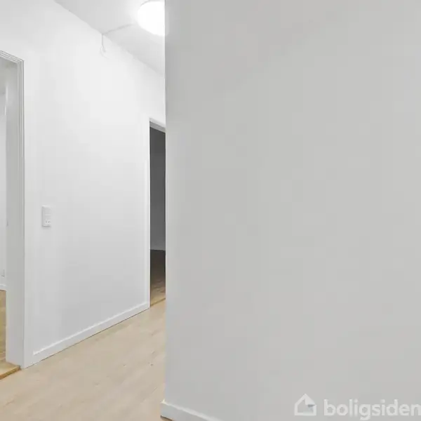 Hvidt rum med åbne døråbninger til venstre. En hvid dørkarm leder til et tilstødende værelse med trægulve. Enkel hvid væg ses til højre.