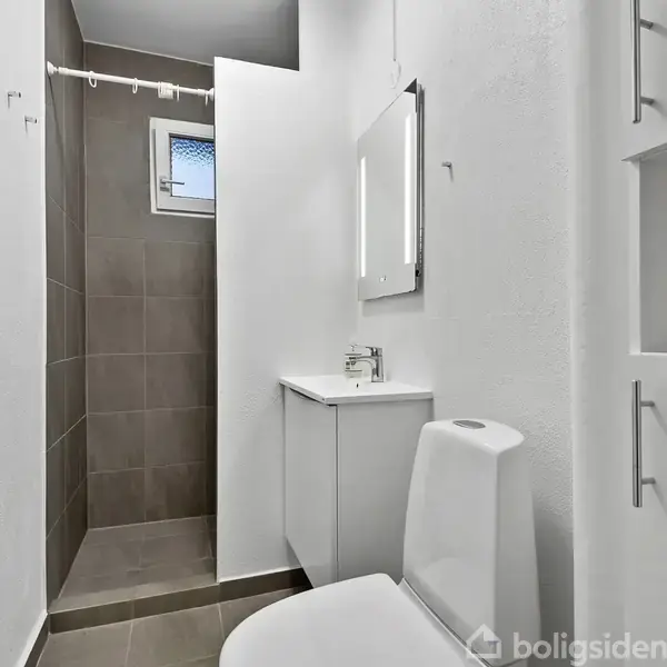 Et badeværelse med brusekabine til venstre, vægmonteret bruseforhæng, vask med spejl i midten og toilet til højre, hvidt interiør med grå fliser.