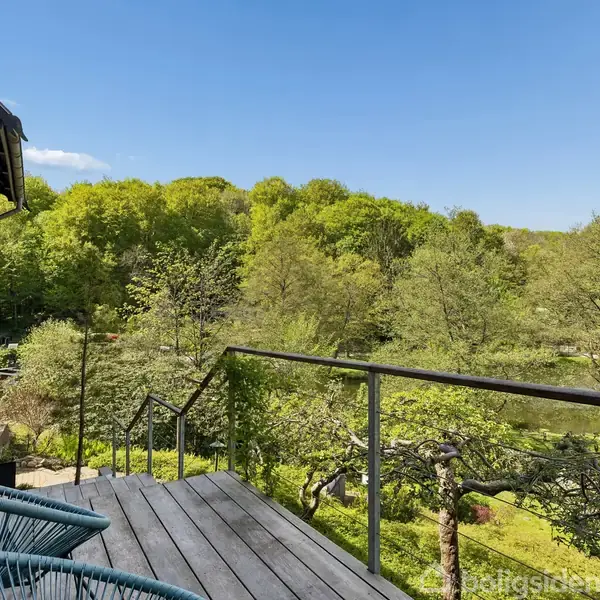 En træterrasse med to moderne stole vender mod en frodig grøn skov i baggrunden, til venstre ses et rødt murstenshus med vinduer, under en klar blå himmel.