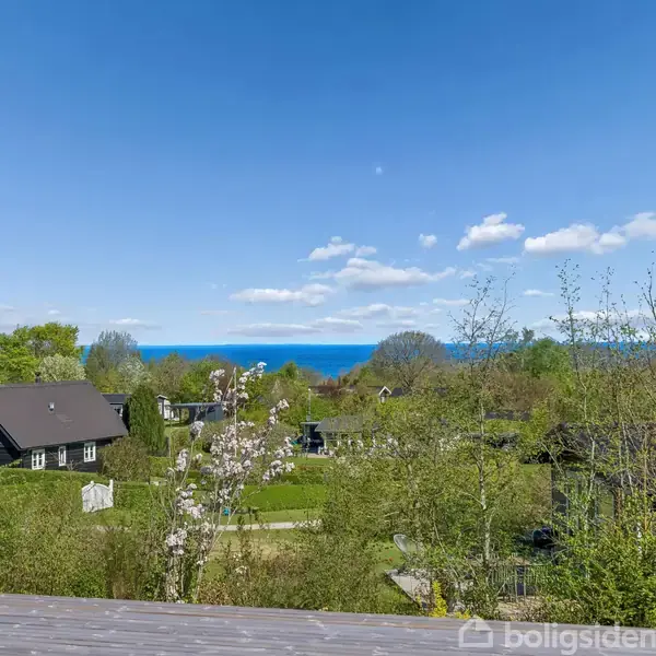 Huse står stille i en frodig, grøn have omkranset af træer. I baggrunden kan havet skimtes under en klar blå himmel med spredte skyer.