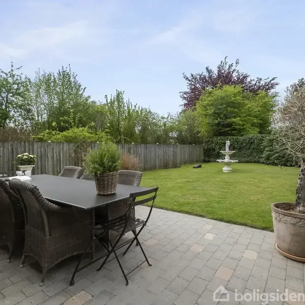 Terrasse med spisebord og stole er omgivet af en grøn have med græsplæne og en hvid fontæne. Et træ i krukke står ved siden, og et hegn omkranser området.