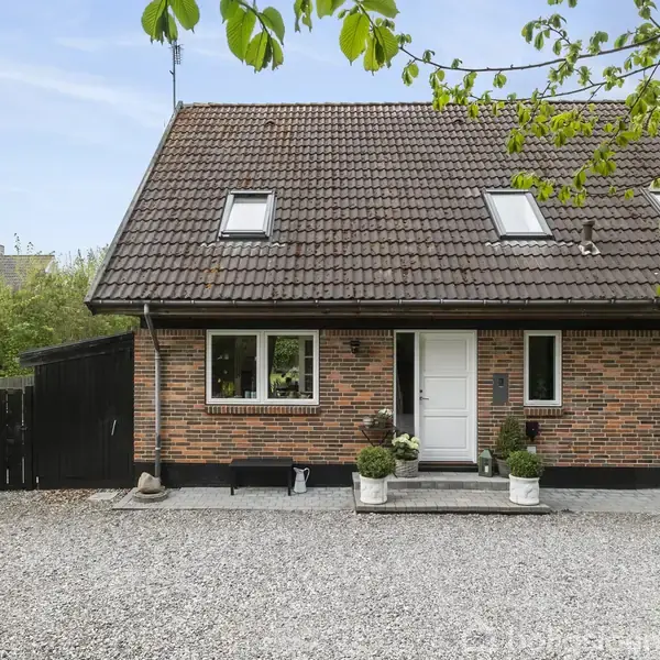 Et rødt murstenshus med brunt tegltag har tre ovenlysvinduer. Indgangen er omgivet af potteplanter. Huset er beliggende i en grusbelagt gårdsplads omgivet af grønne områder.