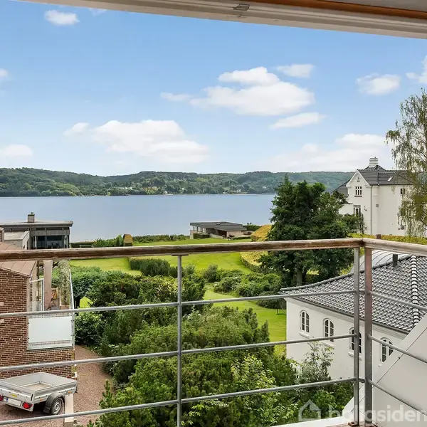 En balkon med gelændere, hvorfra der er udsigt til en sø og frodige grønne områder. I baggrunden ses huse og en klar himmel med få skyer.