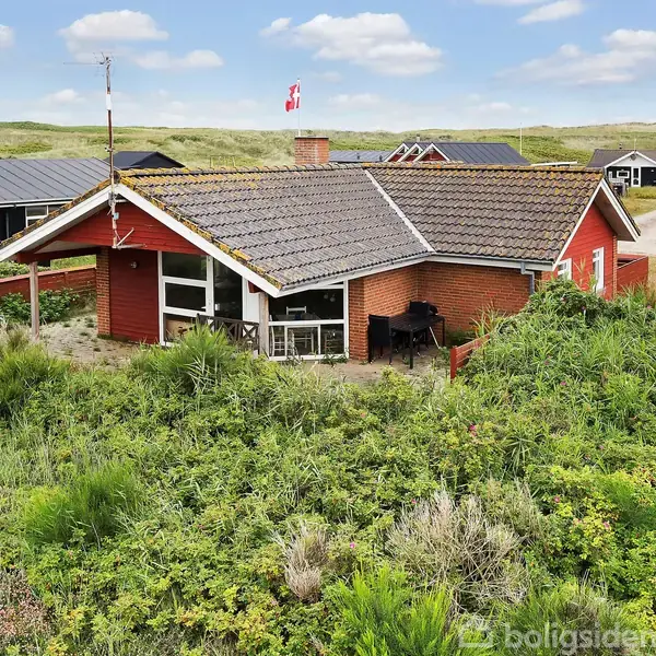 Rødt hus med sort tagflag vajende på en stang; det står omgivet af grønt buskads i et sommerhusområde med flere andre huse i baggrunden under en blå himmel med skyer.