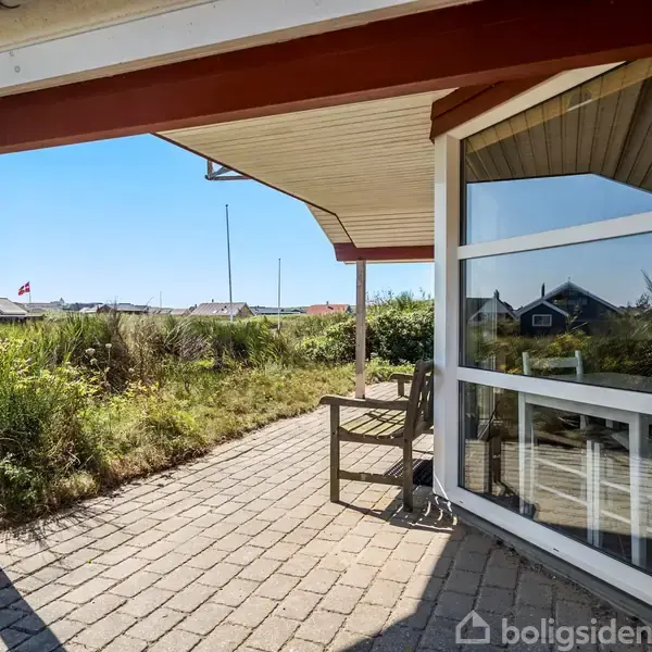 En træterrasse med to stole står under en overdækning. I baggrunden ses græs og blå himmel, hvilket skaber en afslappet, udendørs atmosfære.