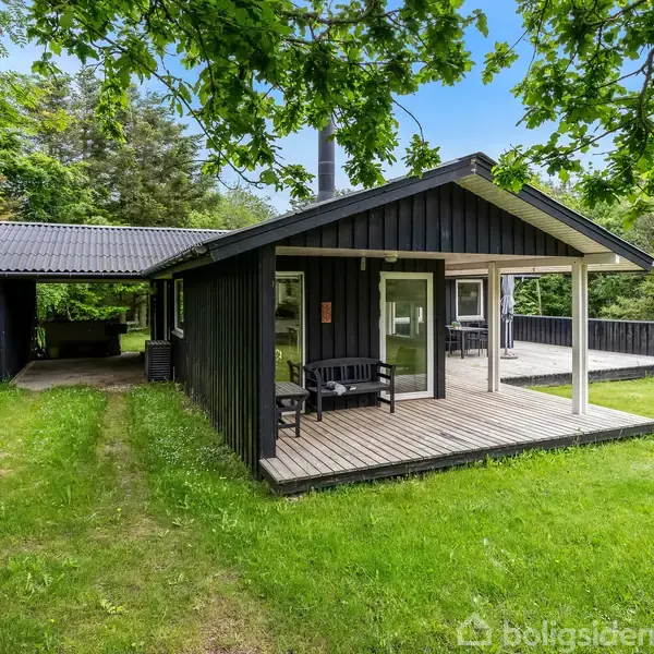 Sort træhus med overdækket terrasse står i en grøn have omgivet af træer. Et bord med to stole er placeret på træterrassen.