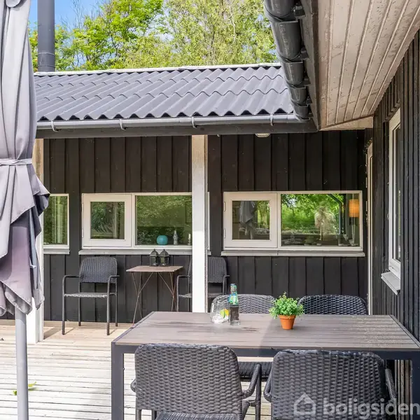 Terrassemøbler står på et trædæk foran et sort, moderne sommerhus. En parasol er foldet sammen i nærheden. Omgivet af grønne træer giver det en rolig atmosfære.