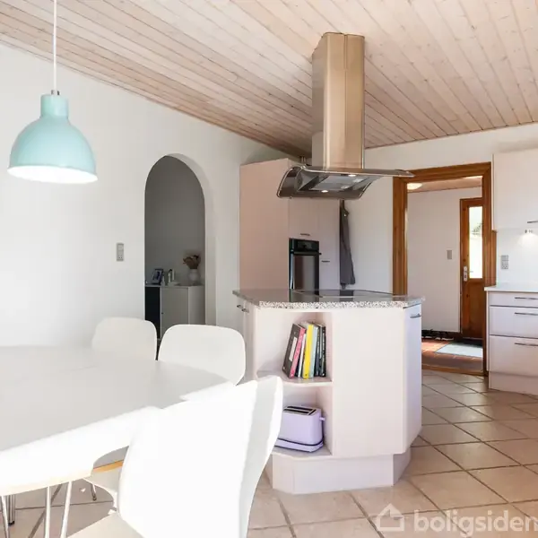 Et rundt, hvidt spisebord med hvide stole står i et lyst køkken. To lyseblå lamper hænger over bordet. Køkkenet har hvide skabe og en åben buegang.