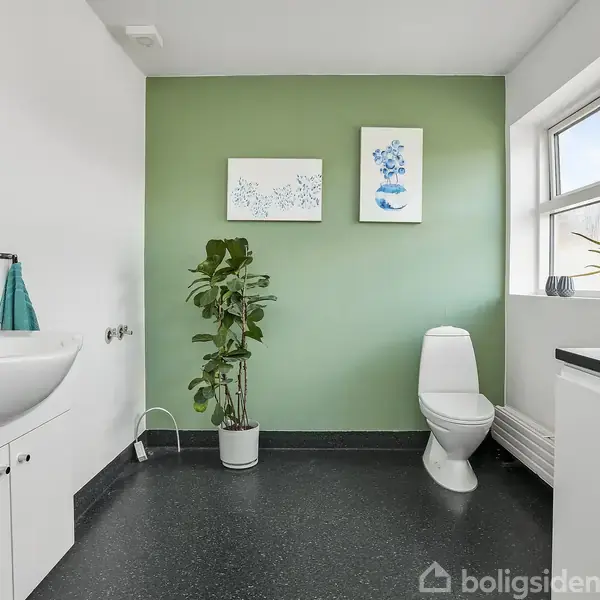 Et badeværelse med en hvid vask og skab til venstre, en grøn plante i midten, og toilet til højre ved en grøn væg. To billeder hænger på væggen.