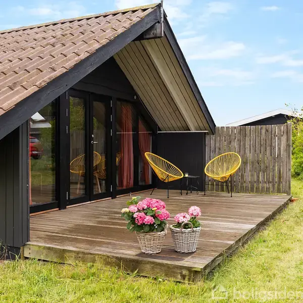 Træhus med store glasdøre åbner ud til en terrasse, hvor to gule stole står. To kurve med lyserøde blomster pynter terrassen. Omgivet af græsplæne og buske i baggrunden.